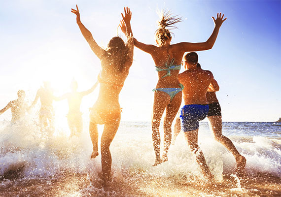 Photo of women in bikinis and men in swim trunks running and jumping with their arms up into the ocean at the beach