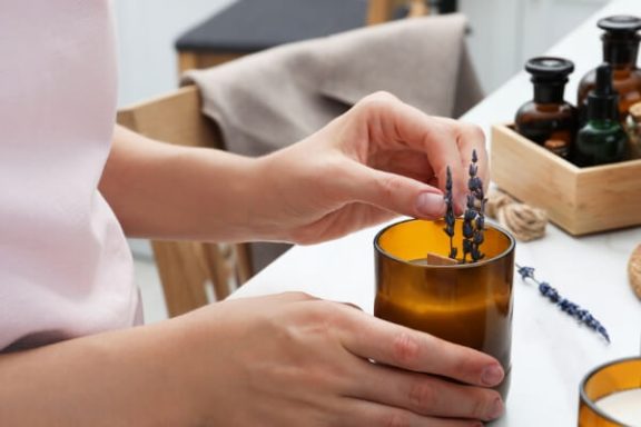 Photo of a woman holding an amber colored candle in her right hand while adding a sprig of lavender to the was with her left hand