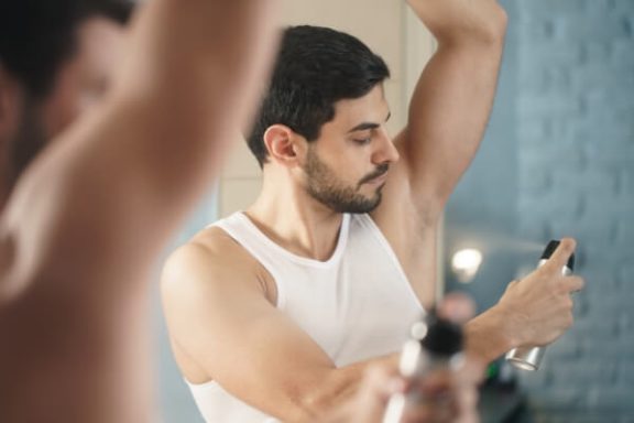 Photo of a man wearing a white tank top spraying deodorant on his underarm out of a can