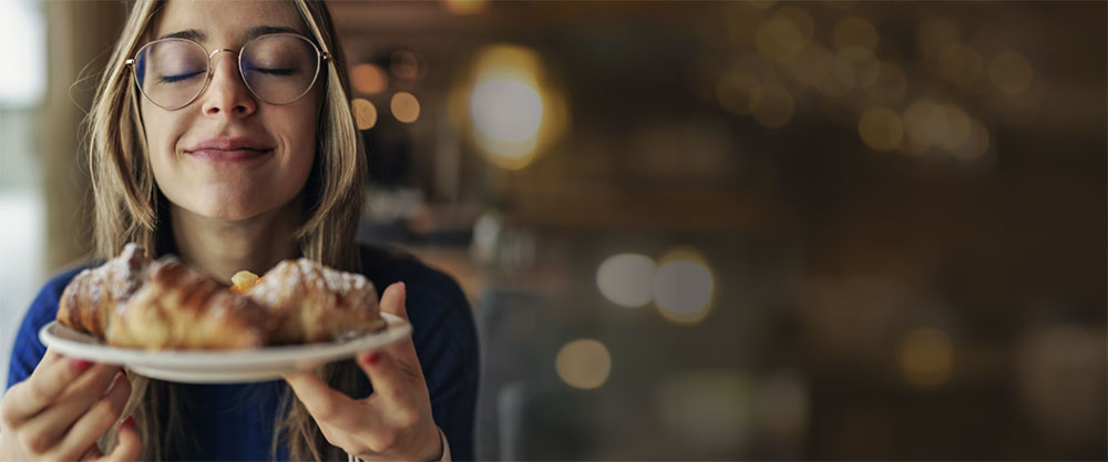 Photo of a woman wearing glasses closing her eyes and smelling the chocolate croissants she is holding on a white plate