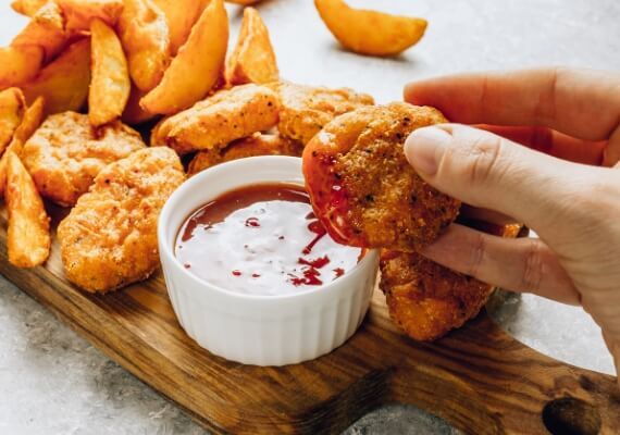 Photo of a chicken nugget that has been dunked in a sauce with more nuggets and potato wedges on a plate