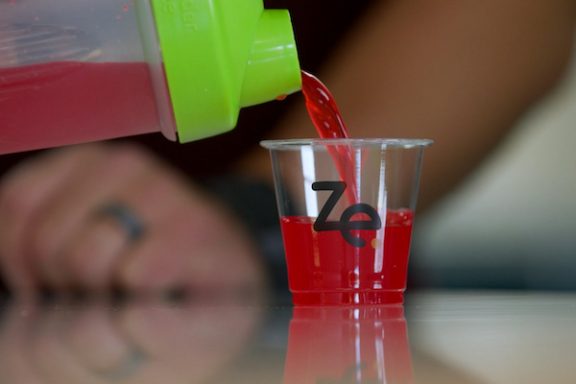Small ZoomEssence cup with the company logo with a red liquid being poured into it