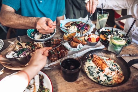 Photo of people reaching with forks to grab food out of a bowl while sitting a table with various dishes in bowls